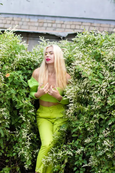 Sarı-yeşil takım elbiseli, uzun saçlı, güzel bir sarışının uzun boylu portresi ve çiçekli yeşil çalıların arka planında parlak yüksek topuklu ayakkabılar.