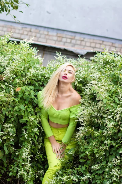 Sarı-yeşil takım elbiseli, uzun saçlı, güzel bir sarışının uzun boylu portresi ve çiçekli yeşil çalıların arka planında parlak yüksek topuklu ayakkabılar.