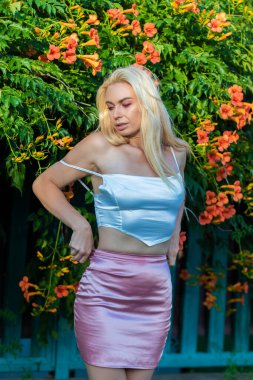 Uzun saçlı, saten beyaz bluz ve pembe etek giyen sarışın bir kadının portresi. Gün batımı. Büyük turuncu çiçeklerle çalıların yanında.