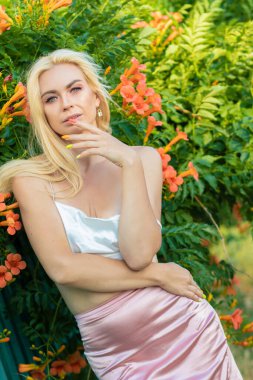 Uzun saçlı, saten beyaz bluz ve pembe etek giyen sarışın bir kadının portresi. Gün batımı. Büyük turuncu çiçekli çalıların olduğu bir çitin yanında