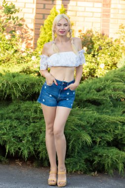 Sarışın kız, kot şortlu, çiçekli ve beyaz bluzlu yeşil bitkilerin arka planına karşı.