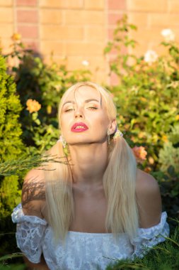 Sarışın kız, kot şortlu, çiçekli ve beyaz bluzlu yeşil bitkilerin arka planına karşı.