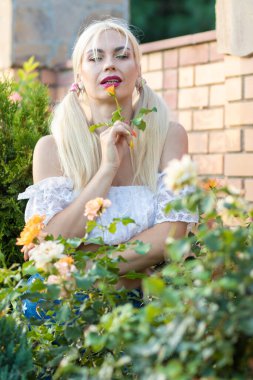 Sarışın kız, kot şortlu, çiçekli, beyaz bluzlu, banliyöde büyük bir tuğla evin arka planında.