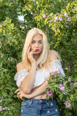 Yeşil bitkilerin arka planında kısa kollu ve kot şortlu dantelli beyaz bir kadının portresi.