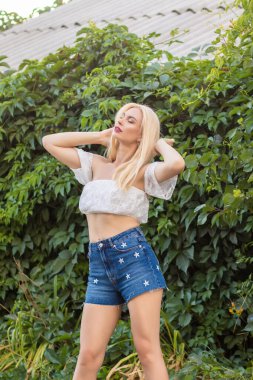 Yeşil bitkilerin arka planında kısa kollu ve kot şortlu dantelli beyaz bir kadının portresi.