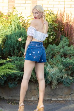 Sarışın kız, kot şortlu, çiçekli ve beyaz bluzlu yeşil bitkilerin arka planına karşı.