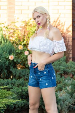 Sarışın kız, kot şortlu, çiçekli ve beyaz bluzlu yeşil bitkilerin arka planına karşı.