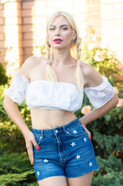 Sarışın kız, kot şortlu, çiçekli ve beyaz bluzlu yeşil bitkilerin arka planına karşı.