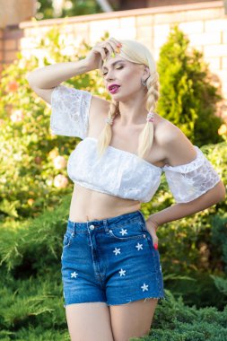 Sarışın kız, kot şortlu, çiçekli ve beyaz bluzlu yeşil bitkilerin arka planına karşı.