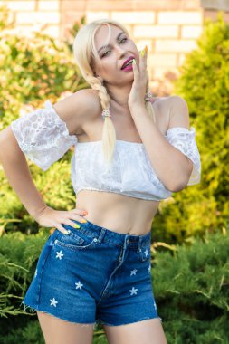 Sarışın kız, kot şortlu, çiçekli ve beyaz bluzlu yeşil bitkilerin arka planına karşı.