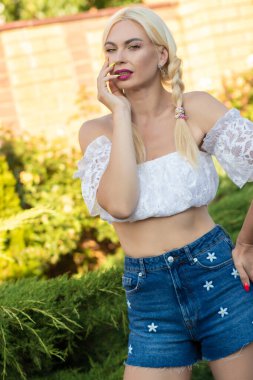 Sarışın kız, kot şortlu, çiçekli ve beyaz bluzlu yeşil bitkilerin arka planına karşı.