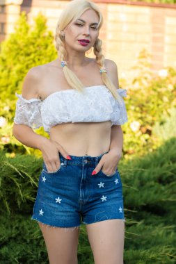 Sarışın kız, kot şortlu, çiçekli ve beyaz bluzlu yeşil bitkilerin arka planına karşı.