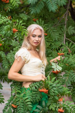 Sarı korseli genç bir kadın, çıplak göbekli ve yeşil bir midi-sarılı etekli, bir Rowan ağacının yanında.