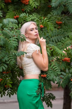 Sarı korseli genç bir kadın, çıplak göbekli ve yeşil bir midi-sarılı etekli, bir Rowan ağacının yanında.