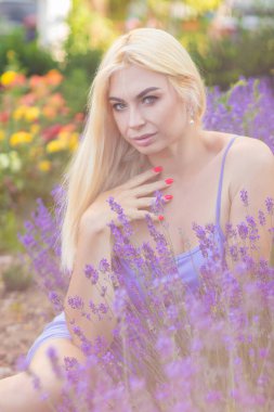 Leylak rengi bir elbisenin içinde uzun saçlı ve doğal makyajlı bir sarışının portresi lavanta çiçeklerinin arasında bir tarlada duruyor. Yaz sabahı