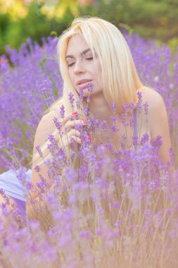 Leylak rengi bir elbisenin içinde uzun saçlı ve doğal makyajlı bir sarışının portresi lavanta çiçeklerinin arasında bir tarlada duruyor. Yaz sabahı