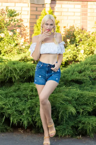 Sarışın kız, kot şortlu, çiçekli ve beyaz bluzlu yeşil bitkilerin arka planına karşı.