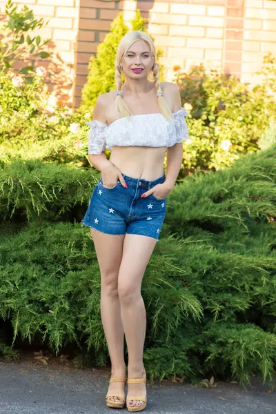 Sarışın kız, kot şortlu, çiçekli ve beyaz bluzlu yeşil bitkilerin arka planına karşı.