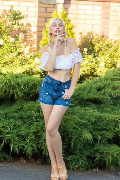 Sarışın kız, kot şortlu, çiçekli ve beyaz bluzlu yeşil bitkilerin arka planına karşı.