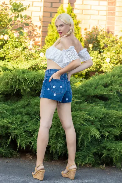 Sarışın kız, kot şortlu, çiçekli ve beyaz bluzlu yeşil bitkilerin arka planına karşı.