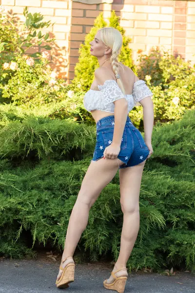 Sarışın kız, kot şortlu, çiçekli ve beyaz bluzlu yeşil bitkilerin arka planına karşı.