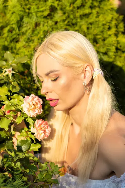 Sarışın kız, kot şortlu, çiçekli ve beyaz bluzlu yeşil bitkilerin arka planına karşı.