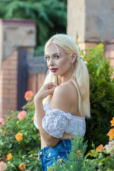 Sarışın kız, kot şortlu, çiçekli, beyaz bluzlu, banliyöde büyük bir tuğla evin arka planında.