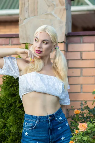 Sarışın kız, kot şortlu, çiçekli, beyaz bluzlu, banliyöde büyük bir tuğla evin arka planında.