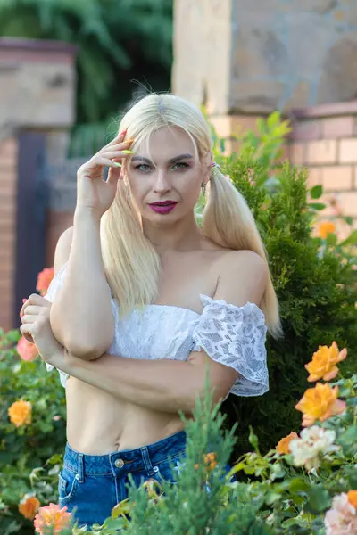 Sarışın kız, kot şortlu, çiçekli, beyaz bluzlu, banliyöde büyük bir tuğla evin arka planında.