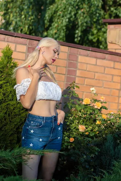 Sarışın kız, kot şortlu, çiçekli, beyaz bluzlu, banliyöde büyük bir tuğla evin arka planında.