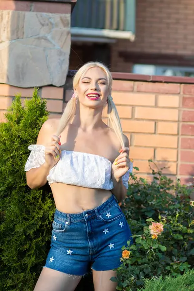 Sarışın kız, kot şortlu, çiçekli, beyaz bluzlu, banliyöde büyük bir tuğla evin arka planında.