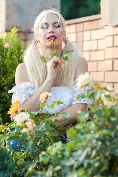 Sarışın kız, kot şortlu, çiçekli, beyaz bluzlu, banliyöde büyük bir tuğla evin arka planında.