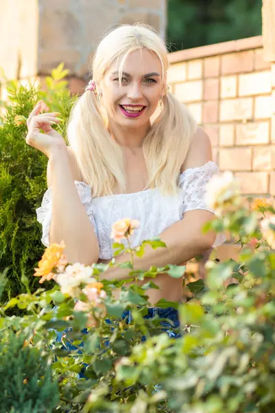 Sarışın kız, kot şortlu, çiçekli, beyaz bluzlu, banliyöde büyük bir tuğla evin arka planında.