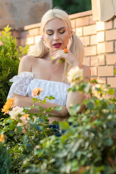 Sarışın kız, kot şortlu, çiçekli, beyaz bluzlu, banliyöde büyük bir tuğla evin arka planında.