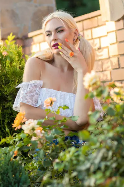 Sarışın kız, kot şortlu, çiçekli, beyaz bluzlu, banliyöde büyük bir tuğla evin arka planında.