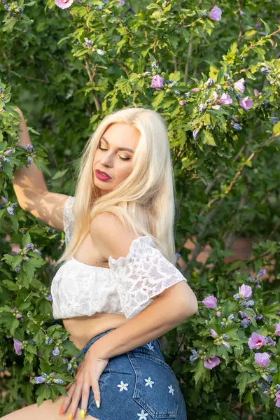 Yeşil bitkilerin arka planında kısa kollu ve kot şortlu dantelli beyaz bir kadının portresi.