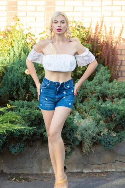 Sarışın kız, kot şortlu, çiçekli ve beyaz bluzlu yeşil bitkilerin arka planına karşı.