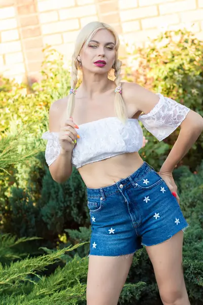 Sarışın kız, kot şortlu, çiçekli ve beyaz bluzlu yeşil bitkilerin arka planına karşı.