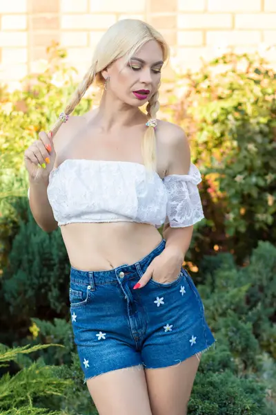 Sarışın kız, kot şortlu, çiçekli ve beyaz bluzlu yeşil bitkilerin arka planına karşı.