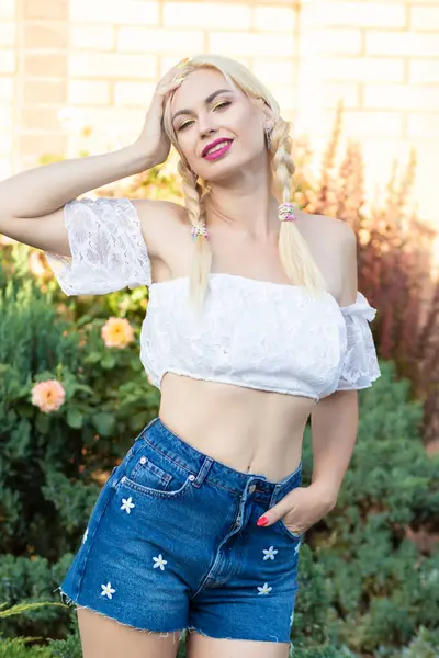 Sarışın kız, kot şortlu, çiçekli ve beyaz bluzlu yeşil bitkilerin arka planına karşı.
