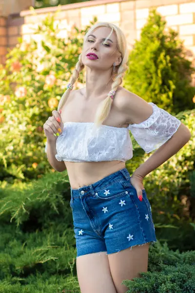 Sarışın kız, kot şortlu, çiçekli ve beyaz bluzlu yeşil bitkilerin arka planına karşı.