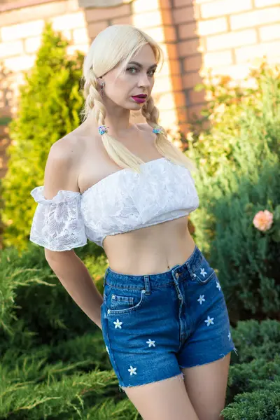 Sarışın kız, kot şortlu, çiçekli ve beyaz bluzlu yeşil bitkilerin arka planına karşı.