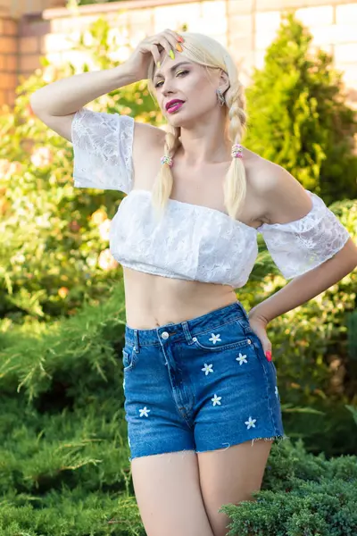 Sarışın kız, kot şortlu, çiçekli ve beyaz bluzlu yeşil bitkilerin arka planına karşı.