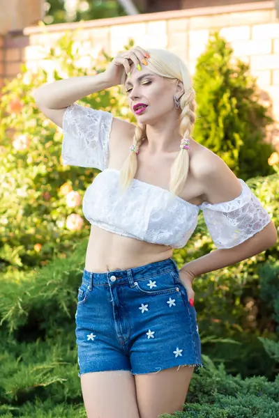 Sarışın kız, kot şortlu, çiçekli ve beyaz bluzlu yeşil bitkilerin arka planına karşı.