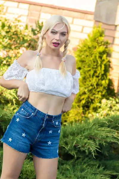 Sarışın kız, kot şortlu, çiçekli ve beyaz bluzlu yeşil bitkilerin arka planına karşı.