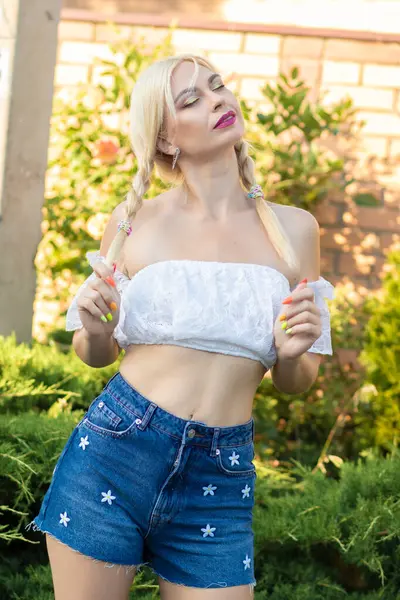 Sarışın kız, kot şortlu, çiçekli ve beyaz bluzlu yeşil bitkilerin arka planına karşı.