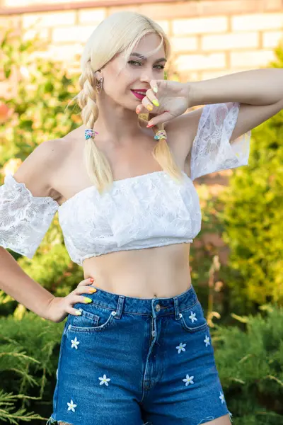 Sarışın kız, kot şortlu, çiçekli ve beyaz bluzlu yeşil bitkilerin arka planına karşı.
