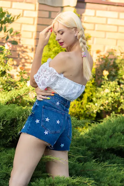 Sarışın kız, kot şortlu, çiçekli ve beyaz bluzlu yeşil bitkilerin arka planına karşı.