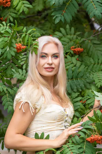 Sarı korseli genç bir kadın, çıplak göbekli ve yeşil bir midi-sarılı etekli, bir Rowan ağacının yanında.