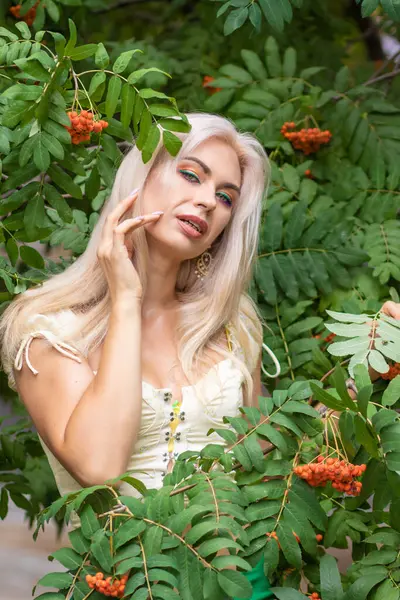 Sarı korseli genç bir kadın, çıplak göbekli ve yeşil bir midi-sarılı etekli, bir Rowan ağacının yanında.